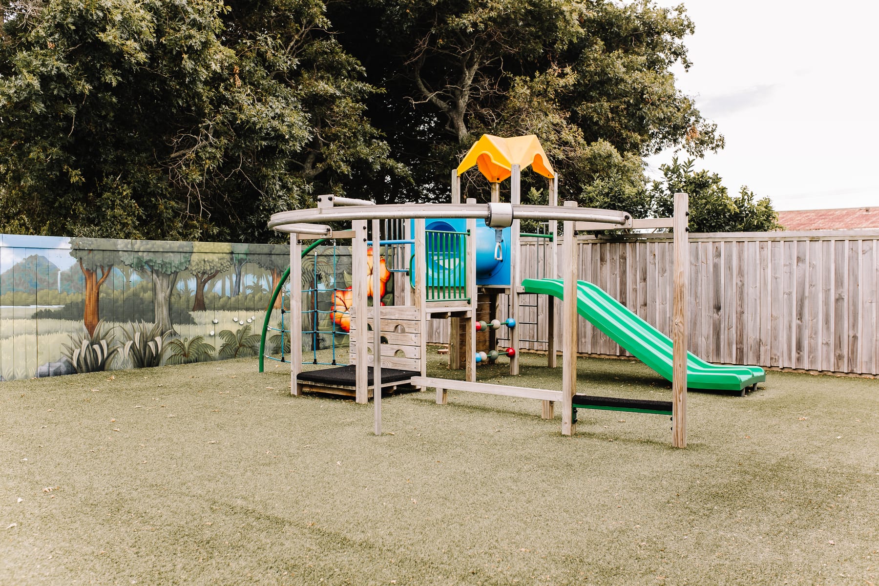 The centre's playground: a wooden climbing frame with a green slide, on grass beneath mature trees.