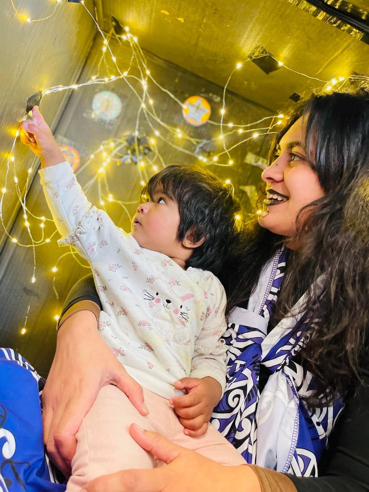 A teacher and a toddler reaching up together to a string of fairy lights indoors.