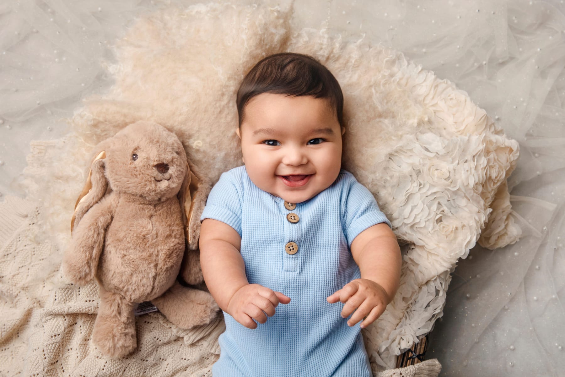 A baby in a pale blue romper lying on a cream fleece, smiling up at the camera beside a soft toy rabbit.