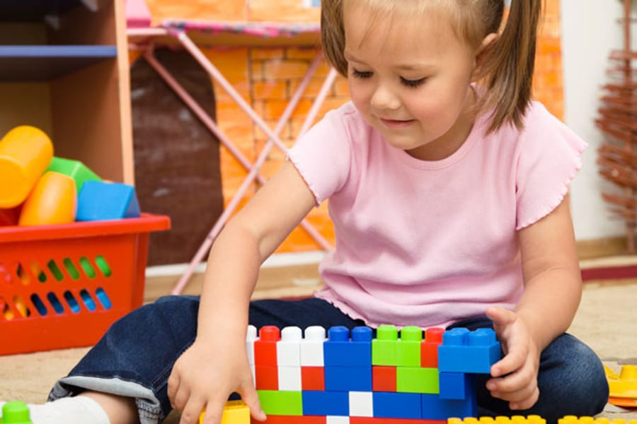 A toddler kneeling on the floor building a low wall out of large plastic bricks.